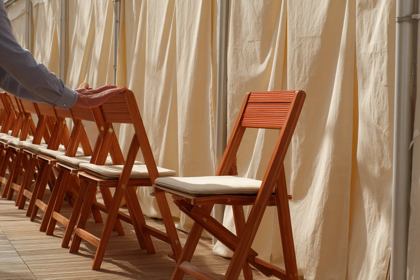 A marquee half set for an event, rows of timber chairs being placed against soft draped fabric in warm daylight.